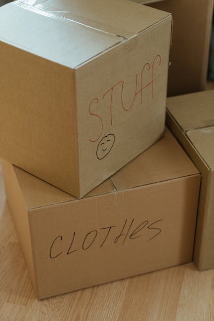 Inside a home during the packing process for a house move, three clearly labeled cardboard boxes are stacked on a wooden floor. The top box has the word 'STUFF' written on it in red marker, accompanie