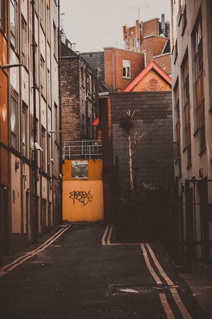 A narrow urban street with tightly packed residential buildings on either side, featuring various architectural styles and materials, including brick and plaster facades. Some buildings have small bal
